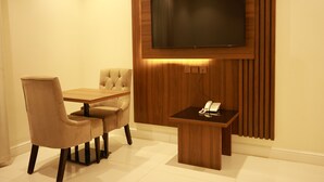 Une chambre moderne dotée d’un grand téléviseur à écran plat fixé sur un mur en panneaux de bois, d’une petite table en bois avec un téléphone et d’un coin repas avec deux chaises beiges à revêtement.