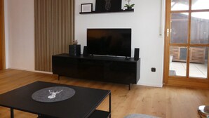 Ein modernes Wohnzimmer mit einem Flachbildfernseher auf einem schwarzen Standfuß, Holzboden, schwarzem Unterhaltungsschrank und einem großen Fenster mit Blick ins Freie.