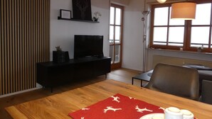Ein modernes Wohnzimmer mit Holzboden, einer schwarzen Unterhaltungseinheit, einem Fernseher, einem Esstisch mit rotem Läufer und einem großen Fenster mit Aussicht.