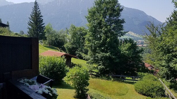 Aussicht auf eine Berglandschaft mit einer Holzbank im Vordergrund, einem Steinhaus und einem klaren Himmel.