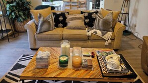 A living room with a beige sofa, patterned cushions, a coffee table with various items, and a kitchen in the background. - Roswell Mini Mansion with Man Cave (Roswell)