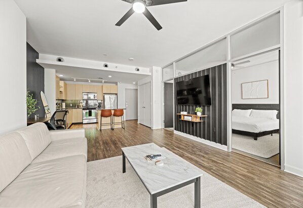 A modern living room with a sofa, a coffee table, and a TV. There is a kitchen in the background with a dining area. - Modern 1-Bedroom City View High Rise Condo with Balcony Gym Pool Free Parking (Dallas)