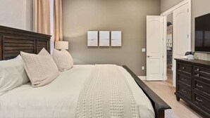 A bedroom with a large bed, a dresser, a TV, and a door leading to another room.