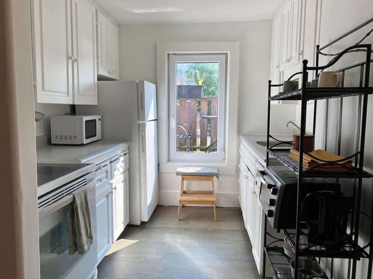Una cocina compacta con armarios blancos, un microondas, un refrigerador, una estufa y un ventanal con vista a un patio trasero.