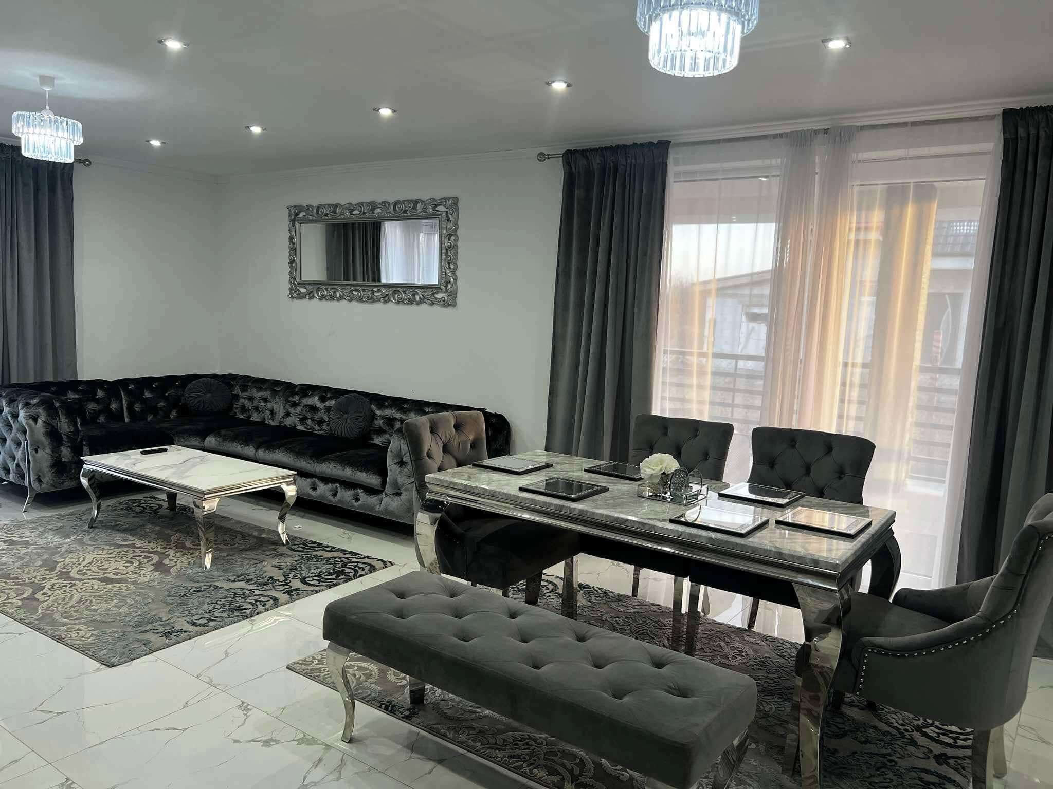 A modern living room with a dark velvet sofa, a glass coffee table, a dining table with chairs, and a chandelier.