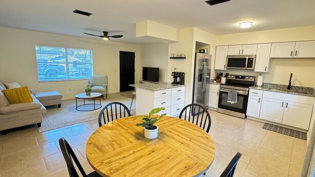 A modern kitchen with white cabinets, a stainless steel refrigerator, and a microwave. There is a dining area with a wooden table and chairs, and a sofa with a yellow pillow. - Charming Lake Town Home with WiFi (Inverness)