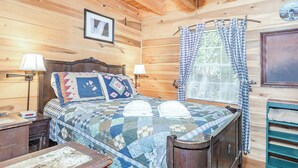 A wooden cabin room with a bed, two bedside lamps, a checkered curtain, and a framed picture on the wall. - Raccoon Loft - Secluded Cabin with Fire Pit and Pickleball (Lake Toxaway)