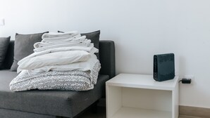 Un lit bien fait, avec des couvertures blanches et à motifs soigneusement pliées, un canapé gris et une table de chevet blanche sur laquelle se trouve un appareil noir.