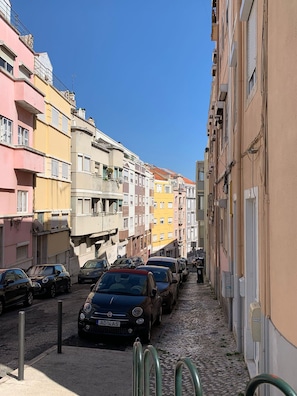 A narrow street lined with colorful buildings, parked cars, and cobblestone pavement.
