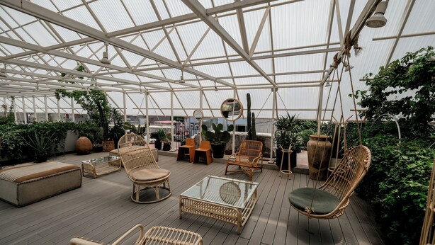 Een ruime kas met een hoge plafond, rieten meubilair en verschillende potplanten.