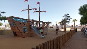 Una estructura de juego de madera con forma de barco pirata, que incluye un tobogán, rodeada de arena y una cerca de madera.