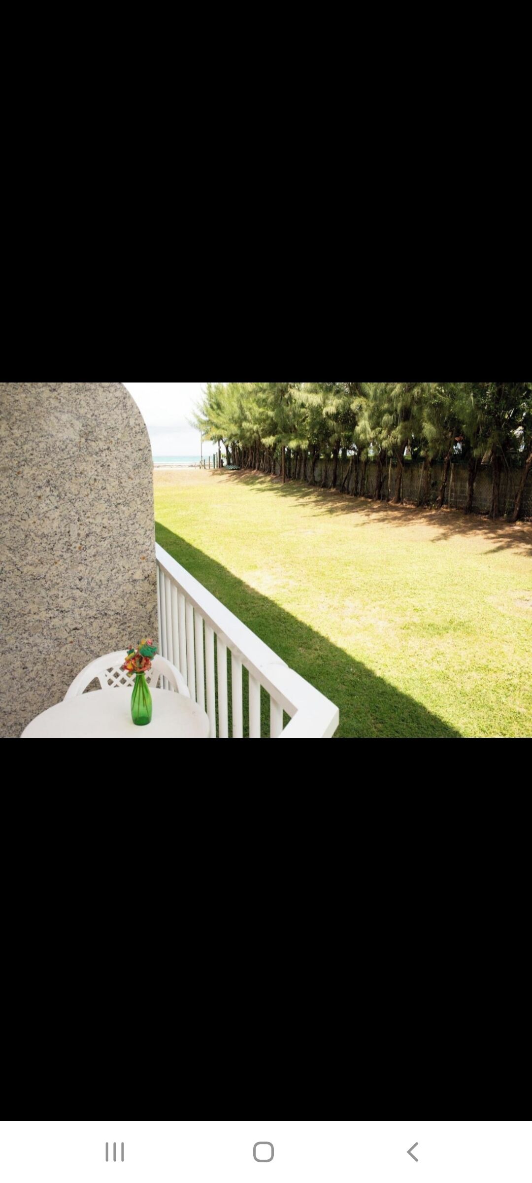 Un balcon avec une balustrade blanche, un vase vert rempli de fleurs et une vue sur un espace verdoyant avec des arbres et une clôture.