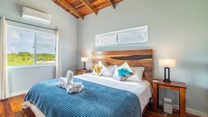 A bedroom with a wooden ceiling, a large bed with a blue bedspread, two bedside tables with lamps, and a window with a view of greenery. - Loggerhead - 2BR House at Secret Beach (San Pedro)
