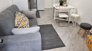 A modern living room with a grey sofa, a small dining table with chairs, a kitchen in the background, and a grey rug. - Light Wave Nazare Center 50m Beach (Nazaré)