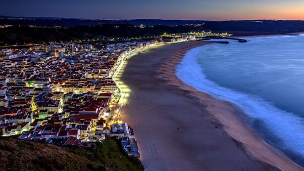 A coastal town at dusk with a beach and illuminated buildings along the shoreline. - Light Wave Nazare Center 50m Beach (Nazaré)