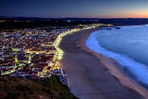 A coastal town at dusk with a beach and illuminated buildings along the shoreline. - Light Wave Nazare Center 50m Beach (Nazaré)