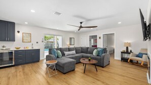 Ein modernes Wohnzimmer mit einem grauen Ecksofa, einem kleinen runden Couchtisch und einem Essbereich mit dunkelgrauen Schränken und einem Einbauofen.