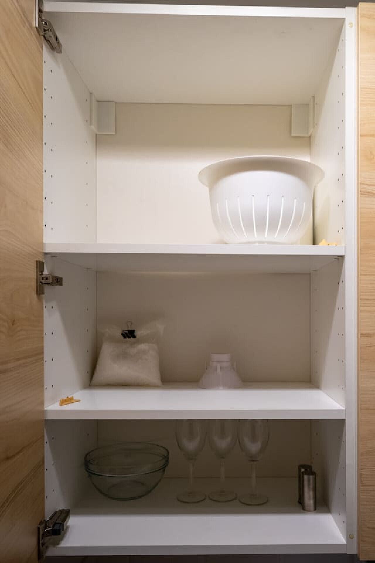 Un armario de cocina blanco con tres estantes; el estante superior sostiene un bol blanco, el estante central tiene una botella blanca y un objeto pequeño, y el estante inferior contiene un bol de vidrio transparente, copas de vino y un recipiente metálico.