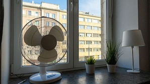 Una habitación con un ventilador de pie, una planta en maceta y una lámpara en el alféizar de la ventana.