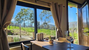 A dining area with a table, chairs, a wine glass, and a view of a green landscape with trees and a fence. - Charming fully equipped cottage in Valle de Guadalupe - Early check in! (Ensenada)
