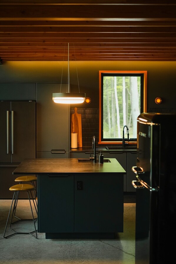 A modern kitchen with a wooden ceiling, a large window with a view of trees, a hanging light fixture, and a black refrigerator. - Black Oak Lodge: A Private Lakeside Retreat (South Frontenac)