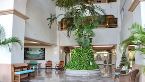 Un hall d’entrée avec un grand jardinier central, des espaces aménagés pour s’asseoir et un tableau accroché au mur.