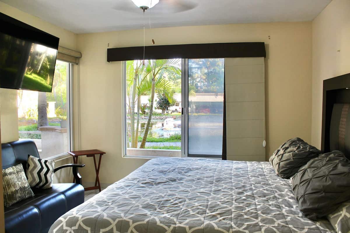 Un dormitorio con cama, una silla, un televisor y una ventana con vistas a zonas verdes y una carretera.