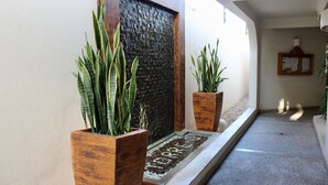 Un couloir avec un mur à texture, deux jardinières en bois contenant des plantes vertes, une petite fontaine décorative et un panneau d’affichage encadré.