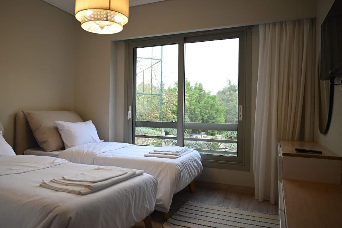 A hotel room with two beds, a large window with curtains, a TV, and a ceiling light.