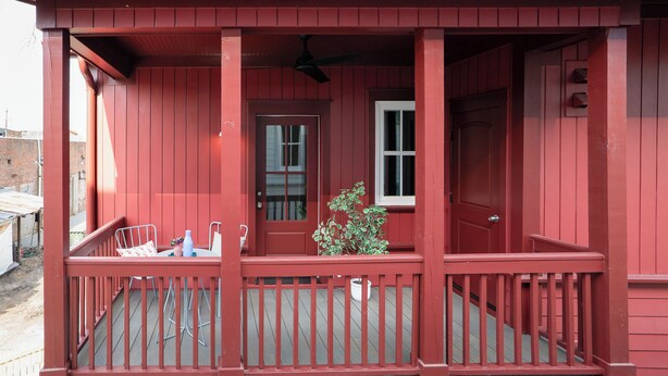 A red wooden porch with a white door, a small potted plant, and a ceiling fan.