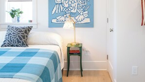 A bedroom with a bed, a blue and white checkered bedspread, a green rug, a wall-mounted lamp, and a large abstract painting.