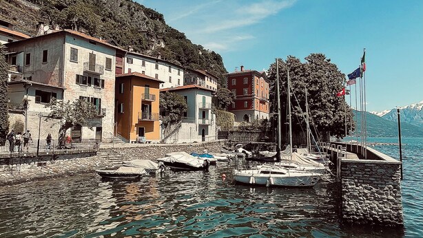 Un village au bord d’un lac, avec des bâtiments qui bordent la rive, un quai où sont amarrés des bateaux et des drapeaux.