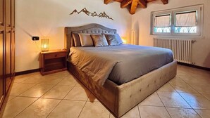 Une chambre avec un grand lit, des poutres apparentes en bois au plafond, une table de chevet avec une lampe, une fenêtre avec des rideaux et une décoration murale représentant des montagnes.