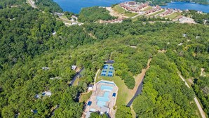 Une vue aérienne d’un complexe hôtelier doté d’une grande piscine, entouré d’une forêt dense, et de plusieurs bâtiments, avec un lac en arrière-plan.