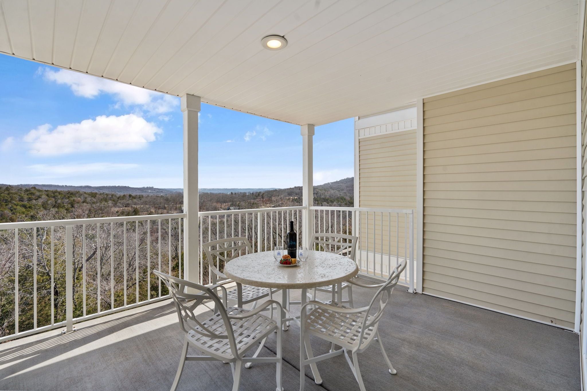 A covered balcony with a round table and chairs, a bottle, and a plate with food.