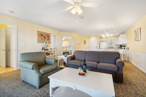 Un salon avec un canapé, un fauteuil et une table basse, un coin repas avec des chaises et une table, et une cuisine à l’arrière-plan.