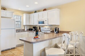 Une cuisine avec des meubles blancs, un îlot central doté de chaises, un réfrigérateur, un four à micro-ondes et une cuisinière.