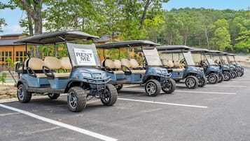 Eine Reihe von Golfcarts auf einem Parkplatz, mit Schildern 'RENT ME', umgeben von Bäumen und einem Gebäude im Hintergrund.