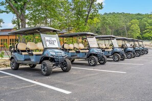 A row of golf carts parked in a lot with 'RENT ME' signs, surrounded by trees and a building in the background.