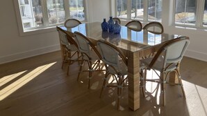 Un comedor con una mesa de vidrio rodeada de sillas de mimbre, ventanas amplias con persianas y vistas al exterior.