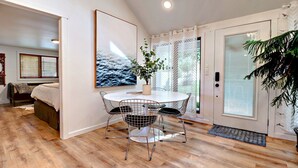 A modern room with a round dining table, metal chairs, a large window with blinds, a bed with a wooden headboard, and a potted plant. - Charming Lakefront Home~Hot Tub~Fire Pit~Large Covered Deck (Fayetteville)