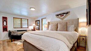 A large bed with a headboard, a bedside table with a lamp, and a window with blinds. - Charming Lakefront Home~Hot Tub~Fire Pit~Large Covered Deck (Fayetteville)