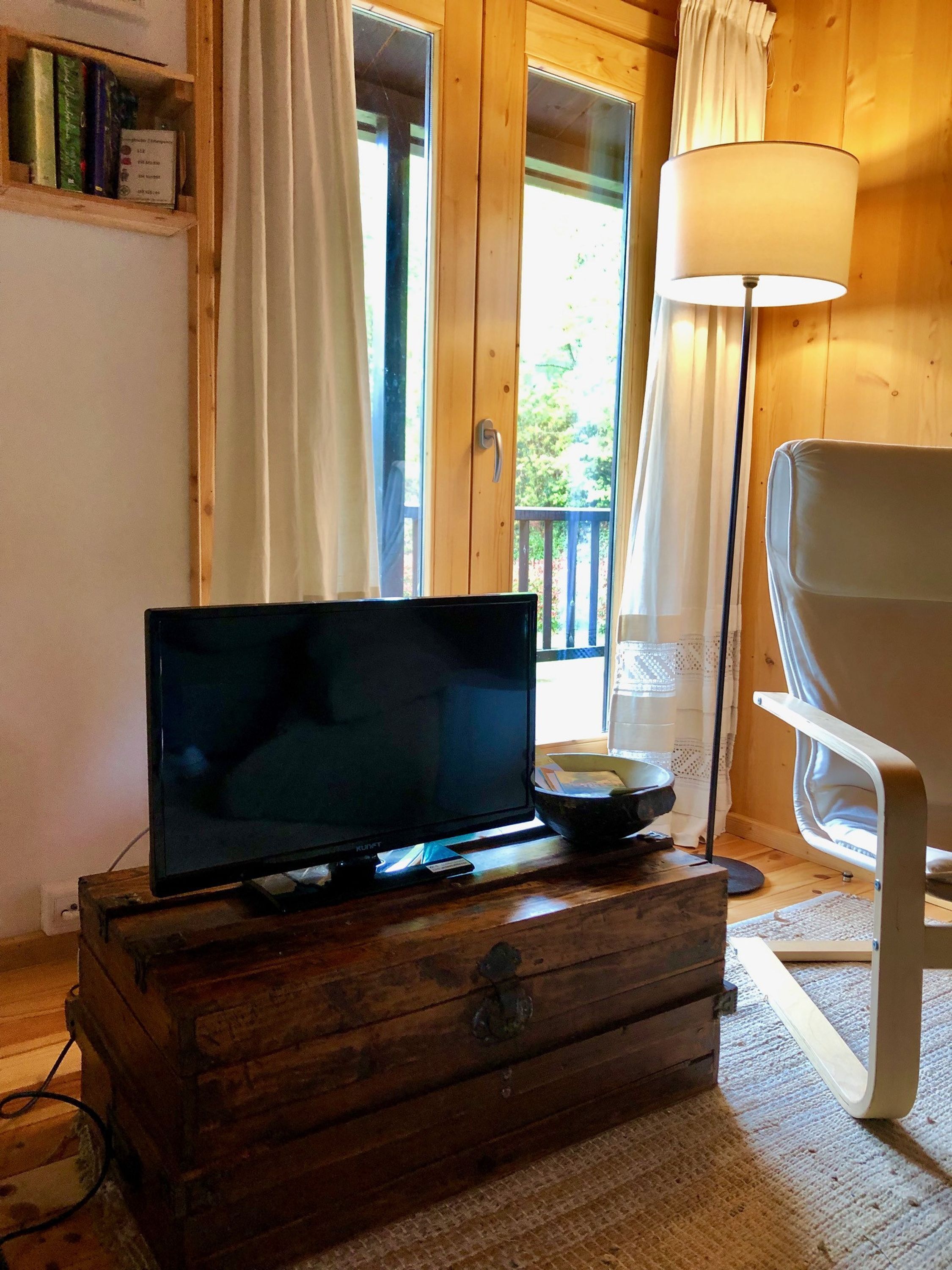 A wooden chest with a television on top, a white chair, a lamp, and a view of the outdoors through a door.