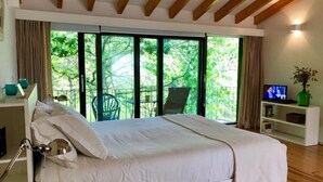 A bedroom with a large bed, a desk with a lamp, a TV, and a view of greenery through large windows.