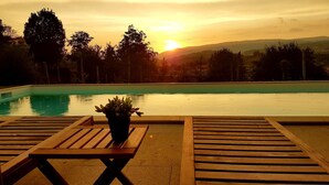 A poolside area with wooden loungers and a potted plant, overlooking a swimming pool and a sunset view.
