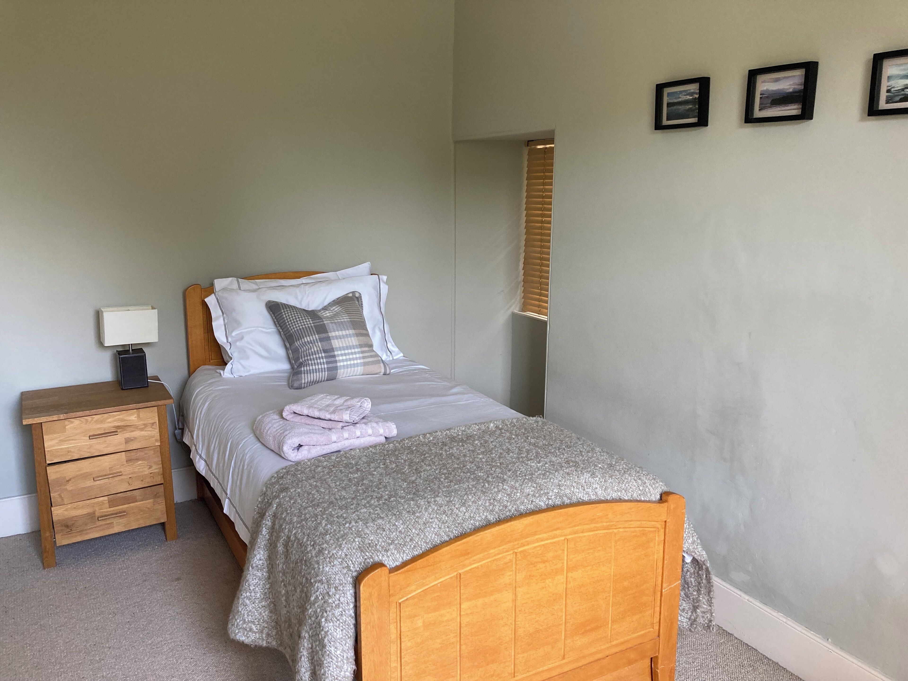 A neatly made bed with a grey blanket and a folded towel, a wooden nightstand with a lamp, and a wall with three framed pictures.