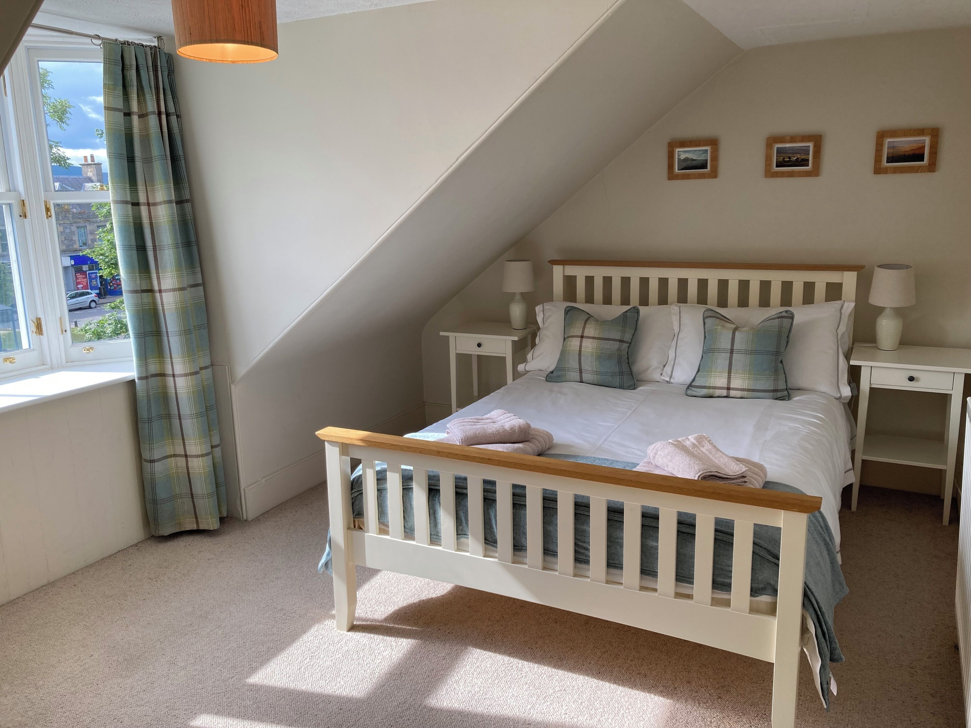 A bedroom with a sloped ceiling, a bed with plaid bedding, two bedside tables with lamps, and a window with curtains.