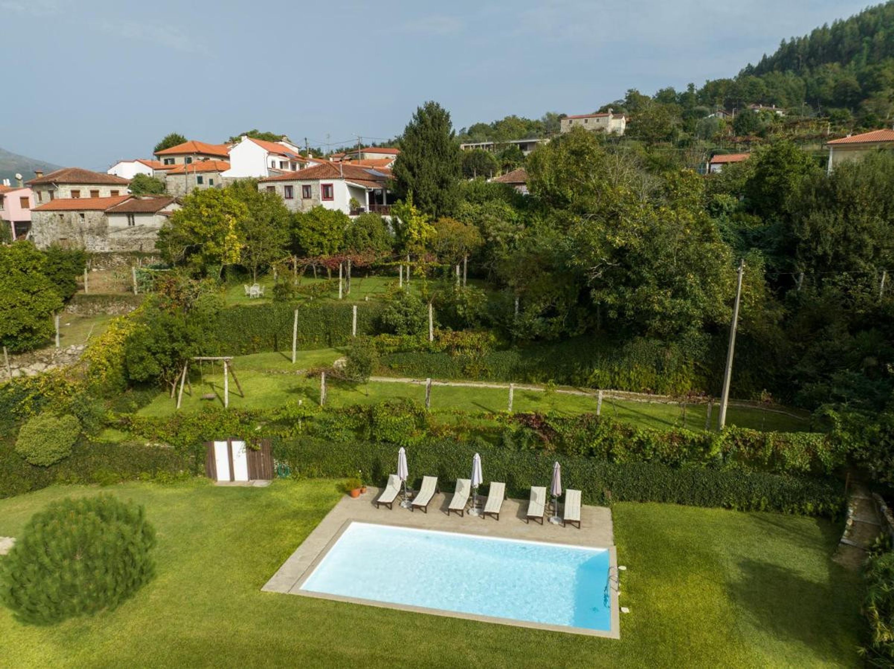 A swimming pool with lounge chairs and a small garden area, surrounded by a hilly landscape with houses in the background.