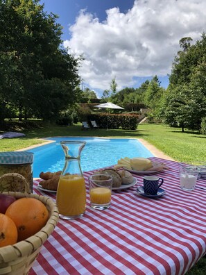 Desayuno junto a la piscina con mantel a cuadros, cesta de frutas, croissants y una jarra de jugo de naranja.