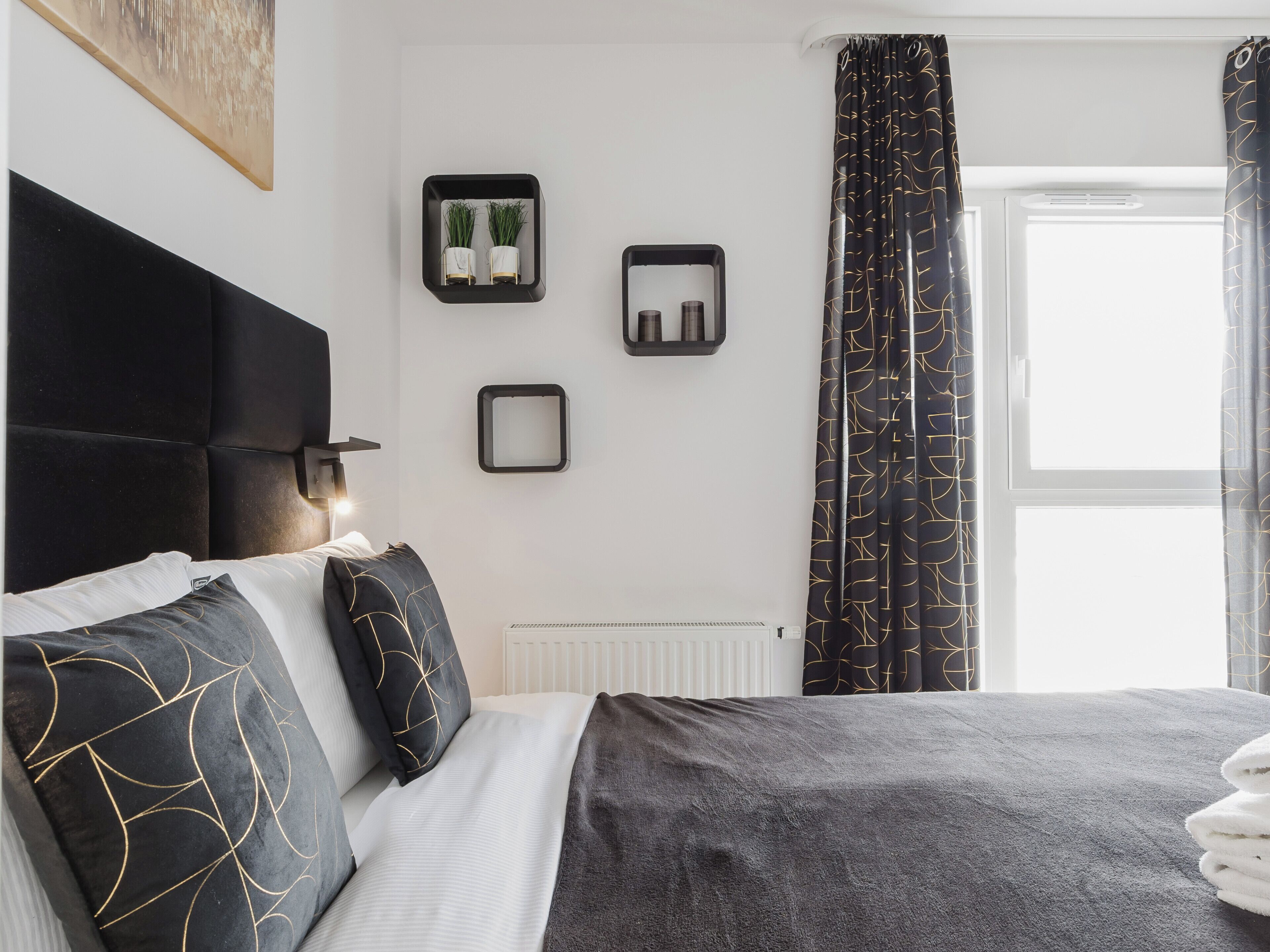 Un dormitorio moderno con cabecera negra, cama con ropa de cama gris y negra, un radiador y una ventana con cortinas estampadas.